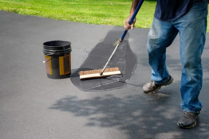 Person painting a dark patch on asphalt driveway with a roller, bucket beside them